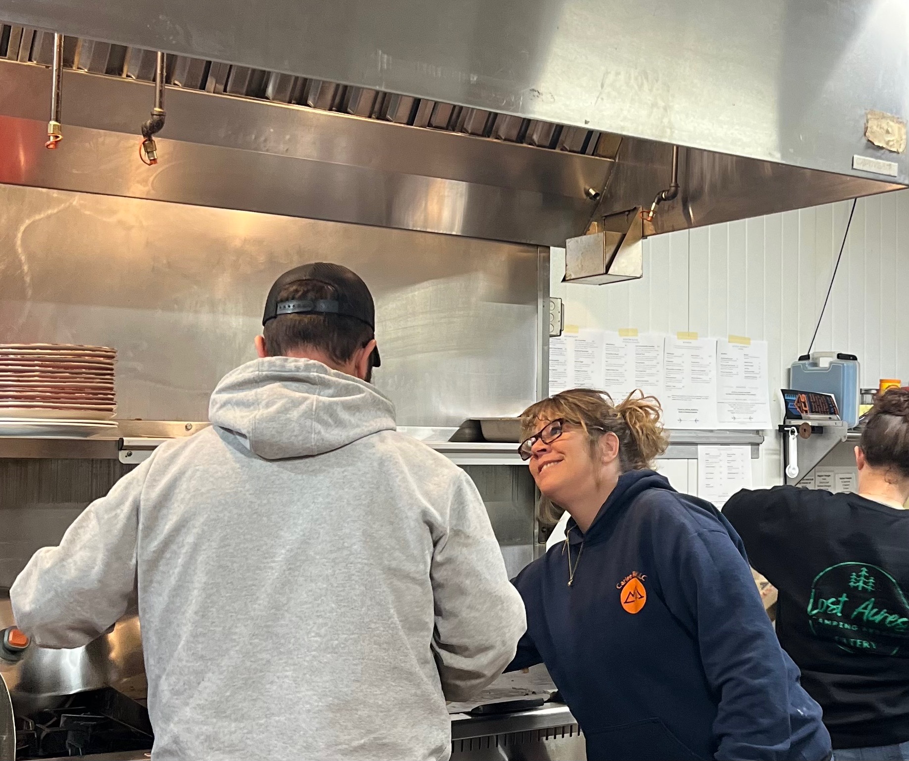 Tracy smiling in the Lost Acres Eatery kitchen beside the line and order tickets