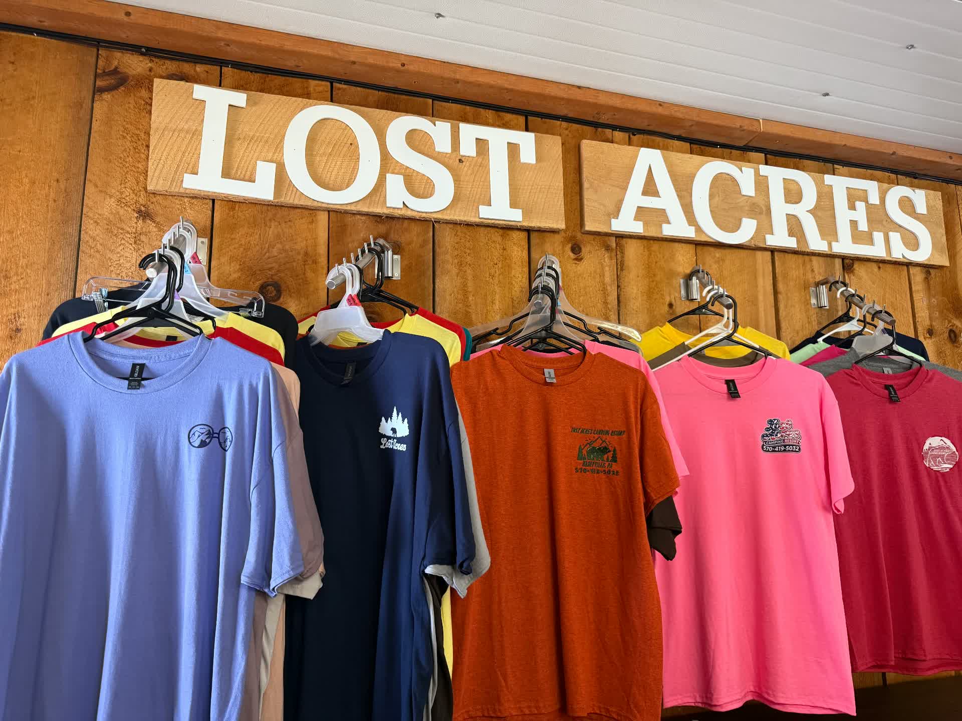 Rack of Lost Acres tees in many colors hanging beneath the wooden LOST ACRES sign